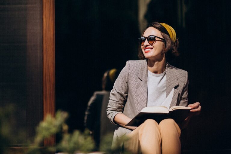 Alt text: American woman sitting outdoors in sunlight, wearing sunglasses, a yellow headband, and a stylish blazer while reading a book and smiling.